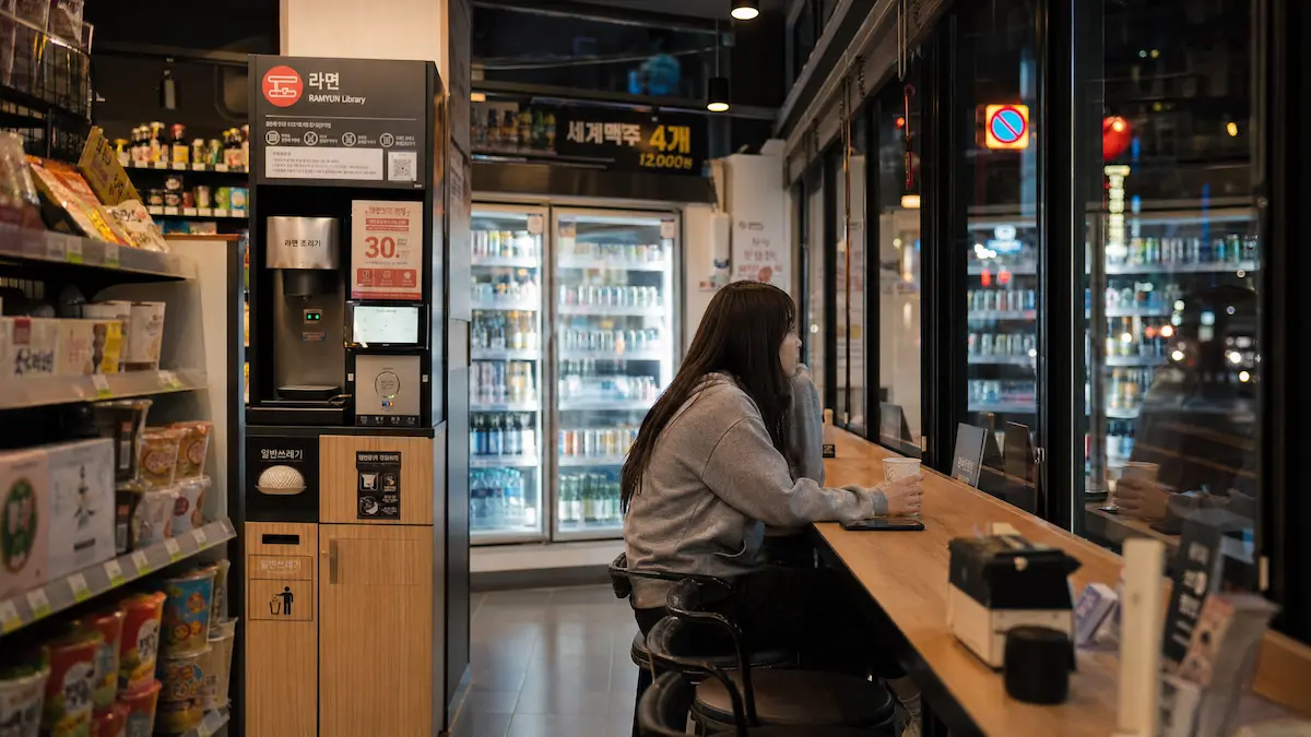 A young woman sitting alone at a counter seat inside a South Korean convenience store late at night, with ramyeon station and glowing refrigerators in the background, illustrating how convenience stores have become a social refuge in modern Korea.