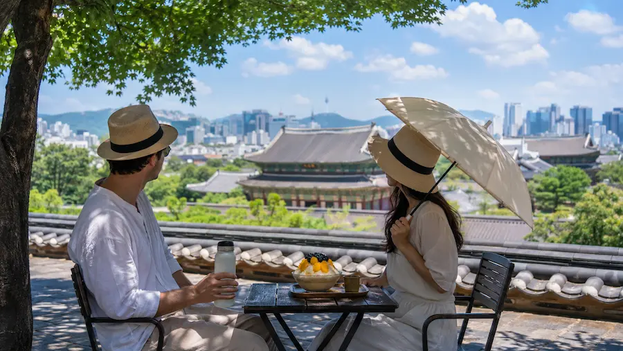 Travelers cooling down with water and bingsu near traditional Korean architecture during a summer heatwave